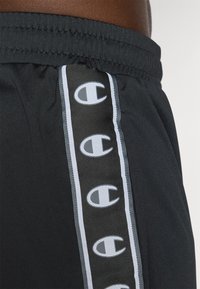 Sorte trening shorts med elastisk livlinje, med en vertikal grå stripe og gjentatte hvite logo aksenter langs siden.