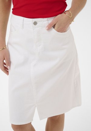 Žena nosi bijelu traper suknju s prednjim prorezom i rukom u džepu, uparenu s crvenom majicom i zlatnim narukvicama na oba zapešća.