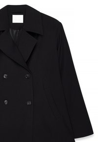 Manteau noir croisé avec larges revers, quatre boutons, poche latérale et doublure intérieure visible sous le col.