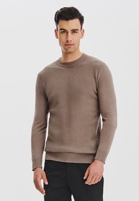 Brązowy sweter z fakturą, okrągłym dekoltem i długimi rękawami, wykonany z dzianiny. Połączony z ciemnymi spodniami.