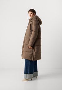 Manteau d'hiver marron avec capuche, présentant un design matelassé, une fermeture éclair et un écusson sur la manche, associé à un jean et des chaussures à talons.