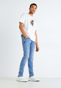 Camiseta blanca de manga corta con estampado gráfico, combinada con jeans de denim azul claro y zapatillas blancas. Diseño simple, ajuste casual.