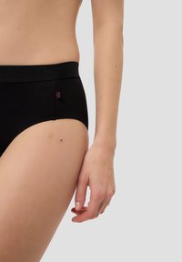 Nahaufnahme einer Person, die schwarze hochgeschnittene Unterwäsche trägt. Der linke Hüfte, die Taille und der entspannte rechte Arm sind gegen einen schlichten Hintergrund zu sehen.