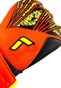 Gant de gardien de but avec un corps orange et des motifs géométriques noirs et jaunes, surface de préhension texturée et sangle de poignet sécurisée.