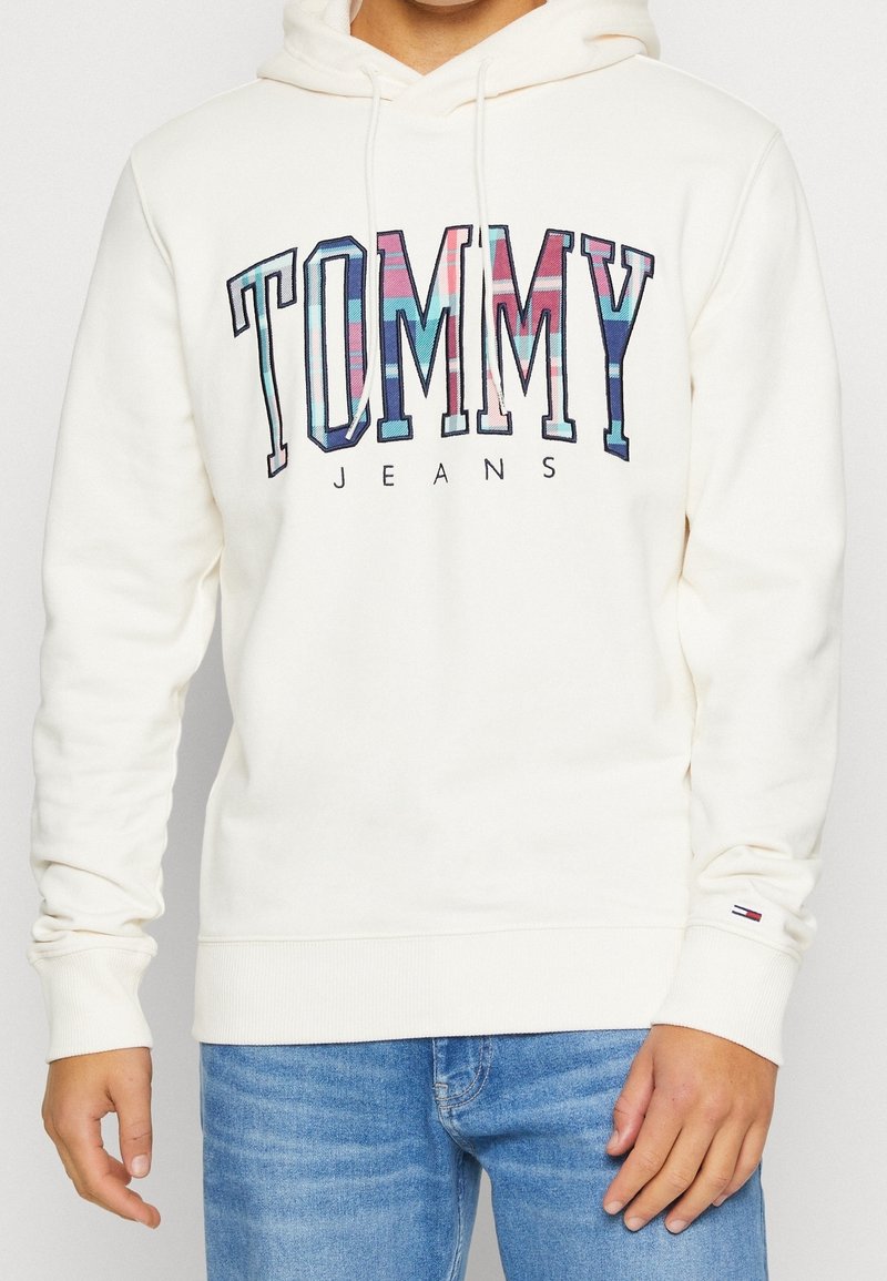 Krémová mikina s kapucí, s multikolorovým kostkovaným logem "TOMMY JEANS". Žebrované manžety a lem, spárovaná se světlými modrými džíny.