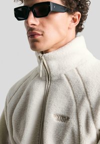 Kremasti dukserica od flisa s visokim ovratnikom i zatvaračem na zip, s pikastim linijama i izvezenim logotipom "UNTITLED GOODS". Nosi crne sunčane naočale.