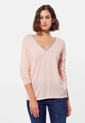 Pull léger rose à col en V avec des manches longues, liseré argenté le long de l'encolure, et coupe décontractée, associé à un jean en denim foncé.