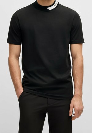 Mann trägt ein schlichtes schwarzes Kurzarm-T-Shirt mit hohem Kragen, das ein schwarz-weißes Design und den Schriftzug „BOSS“ zeigt, kombiniert mit schwarzen Hosen.