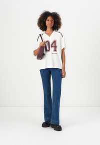 Weißes oversized Hemd mit burgunderfarbener Grafik "04" und MODSTRÖM-Text, kombiniert mit weitem blauen Jeans und schwarzen Plateauschuhen.