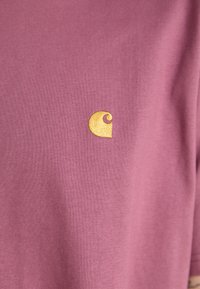 Short-sleeve, crewneck t-shirt in muted pink fabric with a soft texture. Features a prominent golden logo on the chest.