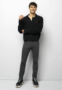 Jonge man die tegen een witte muur staat, gekleed in een zwarte trui met een rood hartembleem, grijze broek en zwart, wit en rode sneakers.