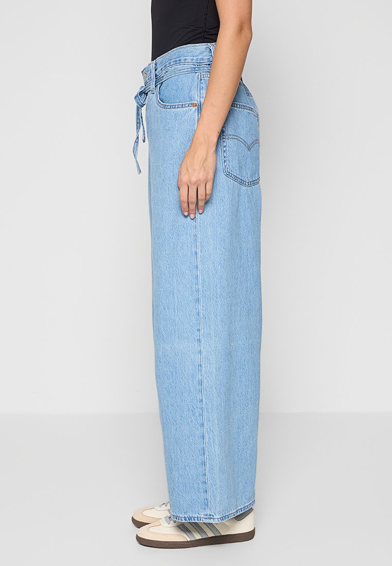Jeans en denim bleu clair taille haute avec une coupe décontractée, dotés d'une ceinture à nouer et de poches latérales. Portés avec des baskets crème.