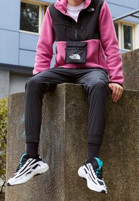 Chaqueta de forro polar negra y rosa, pantalones negros y zapatillas deportivas en negro y blanco con detalles en verde azulado. Modelo sentado sobre una superficie de piedra al aire libre.