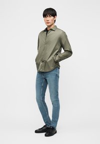 Jeune homme debout les mains dans les poches, portant une chemise à boutons vert olive, un jean slim bleu clair et des baskets noires sur un fond blanc.