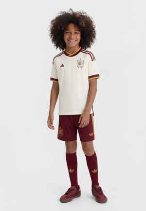 Šypsantis vaikas su garbanotais plaukais, vilkintis balta ir bordo Ispanijos futbolo aprangą, stovintis priešais paprastą šviesią foną.