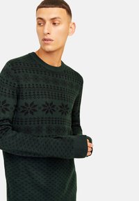 Dark green knitted sweater with black geometric patterns and snowflake designs. Crew neckline and ribbed cuffs. Soft texture.