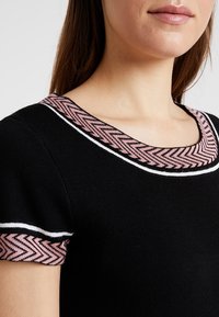 Černý pletený top s kulatým výstřihem, který má růžové herringbone akcenty a bílé lemování na rukávech. Detailní pohled zdůrazňující designové prvky.