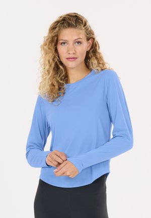 Femme aux cheveux blonds bouclés portant une chemise bleu clair à manches longues et un pantalon noir, debout avec les mains jointes devant.