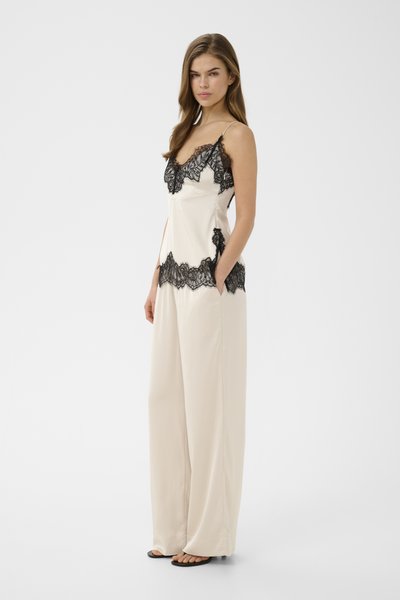 Femme aux longs cheveux porte un pantalon ample crème et un top caraco assorti avec des détails en dentelle noire, mains dans les poches, fond neutre.