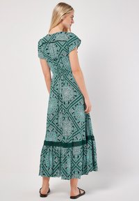 Maxikleid in Grün mit detailliertem Muster. Es hat kurze Ärmel, eine taillierte Taille und einen mehrfach gestuften Saum mit Spitzelementen.