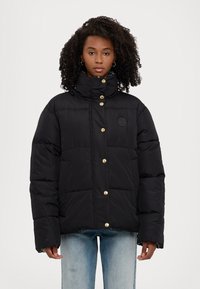 Doudoune noire à col montant, boutons dorés et motif texturé ; associée à un jeans en denim clair.