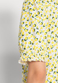 Krótki rękaw z elastycznym mankietem na białym materiale w małe żółte cytryny i zielone liście, noszony na ramieniu.