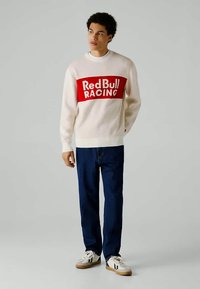 Kremasti pulover s rdečim pasom, na katerem piše "Red Bull RACING." V kombinaciji s temno modrimi kavbojkami in belimi supergami s smešnimi rjavimi podplati.