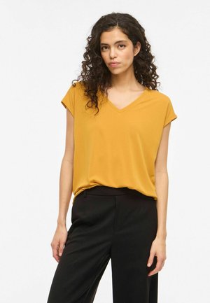 Femme aux cheveux bouclés et foncés, portant un haut jaune moutarde à manches courtes et col en V, ainsi qu'un pantalon noir, debout devant un fond blanc uni.