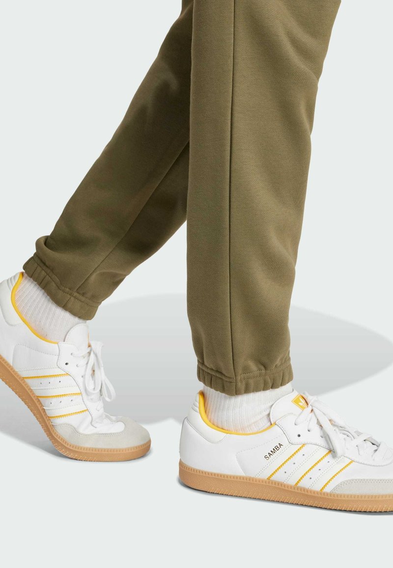 Osoba nosząca białe sneakersy Adidas Samba z żółtymi akcentami oraz oliwkowe spodnie dresowe, stojąca na jasnym tle.