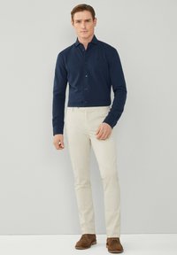 Camicia blu navy con bottoni e maniche lunghe, abbinata a pantaloni crema e scarpe in suede marrone. Tessuto strutturato, design attillato e colori a contrasto.