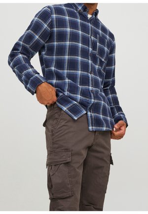 Man draagt blauw geruit overhemd en bruine cargobroek, terwijl hij met één hand de zoom van zijn overhemd recht trekt en met de andere hand in zijn zak zit.