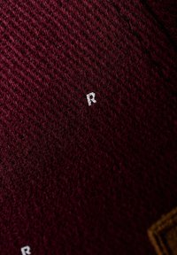 Tejido de punto burdeos con un patrón texturizado, que presenta un pequeño detalle bordado en blanco con la letra "R" en la superficie.