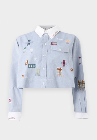 Camisa corta azul claro con cuello y puños blancos, decorada con parches bordados de colores y un bolsillo en el pecho con solapa abotonada.