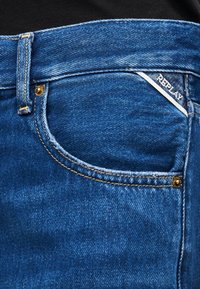 Κοντινό πλάνο τσέπης από μπλε τζιν με ορατές ραφές, χρυσά καρφιά και μια μικρή λευκή ετικέτα "REPLAY" σε μαύρο φόντο.