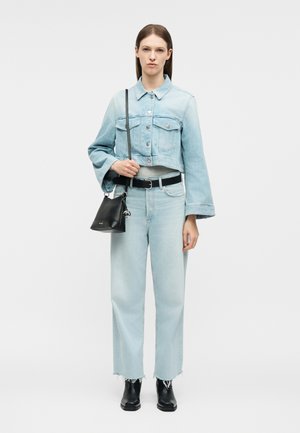 Γυναίκα που φοράει ανοιχτό μπλε τζιν τζάκετ cropped και τζιν παντελόνι, μαύρη ζώνη και μπότες, κρατώντας μαύρη τσάντα ώμου με λογότυπο.