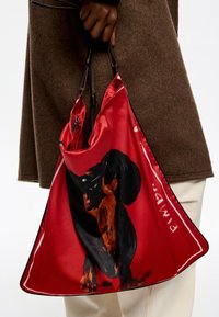 Czerwona satynowa torba na ramię z ilustracją czarno-brązowego psa, z ciemnymi skórzanymi paskami i czarnym wykończeniem na krawędzi.