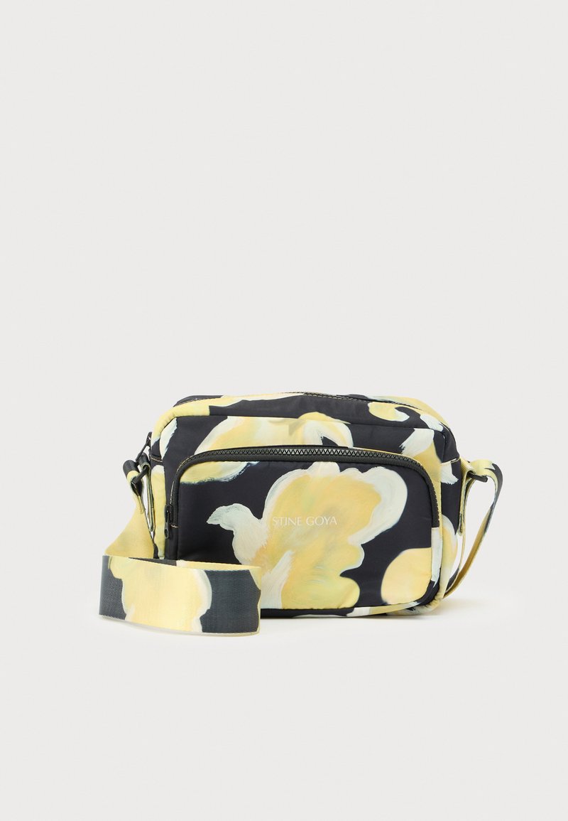 Black fabric bag with large yellow floral pattern, featuring a zippered front pocket and adjustable strap. Stine Goya branding visible.