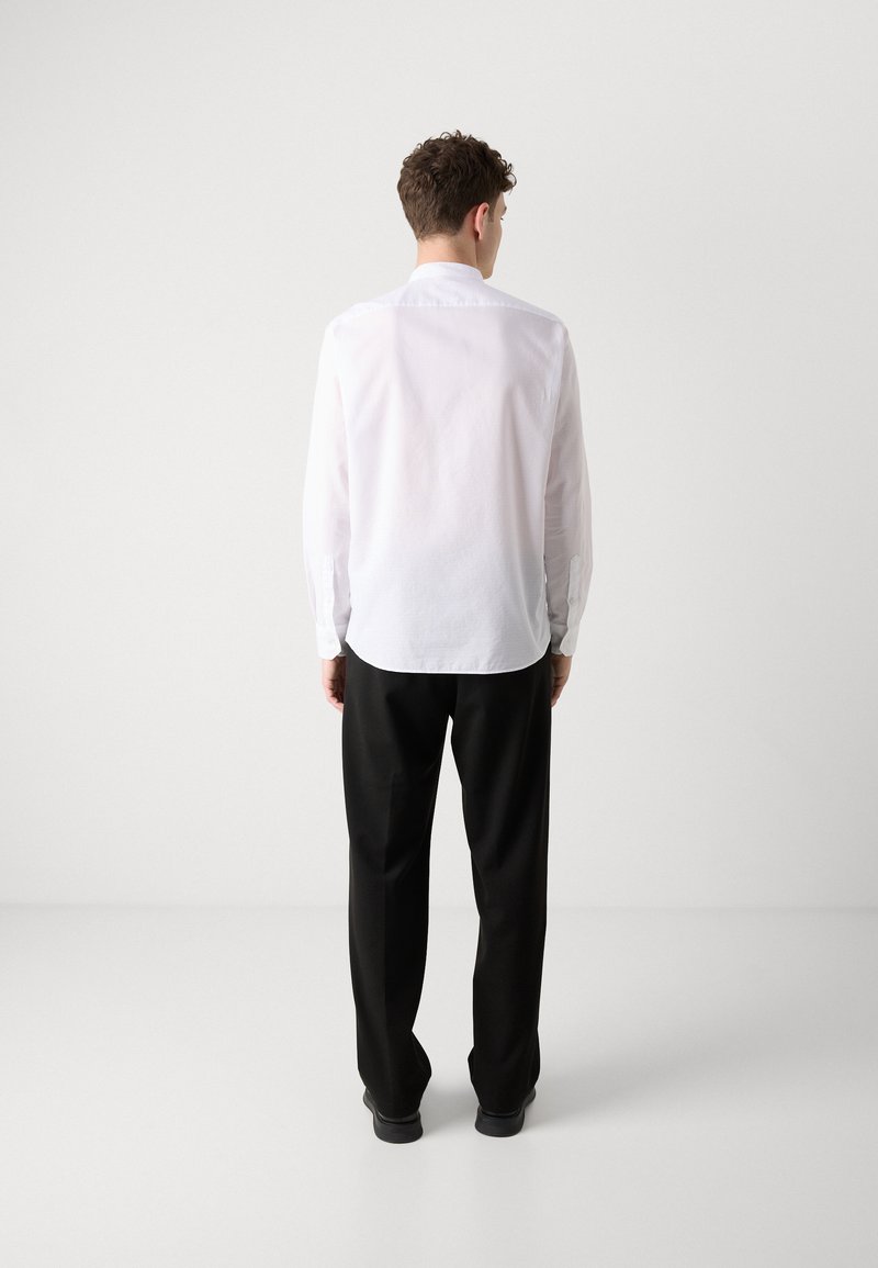 Biała koszula z długim rękawem i guzikami przy mankietach, o gładkiej fakturze i zaokrąglonym dole, w połączeniu z czarnymi spodniami i płaskimi butami.