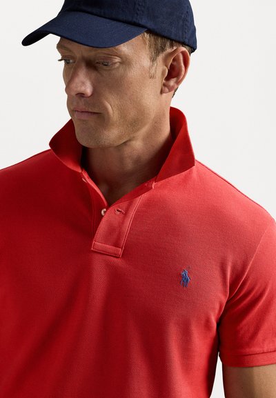 Röd polo t-shirt med en texturerad tyg, med en blå broderad logotyp på bröstet och en upphöjd krage. Bärs med en marinblå keps.