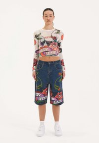 Blusa blanca de manga larga con coloridos diseños de tatuajes florales, combinada con pantalones cortos de denim que presentan gráficos florales similares y acentos de texto. Zapatillas blancas.