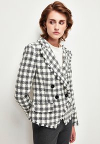 Blazer a cuadros en gris y blanco con diseño de doble botonadura, bordes deshilachados, botones negros y dos bolsillos delanteros. Ajuste estructurado.