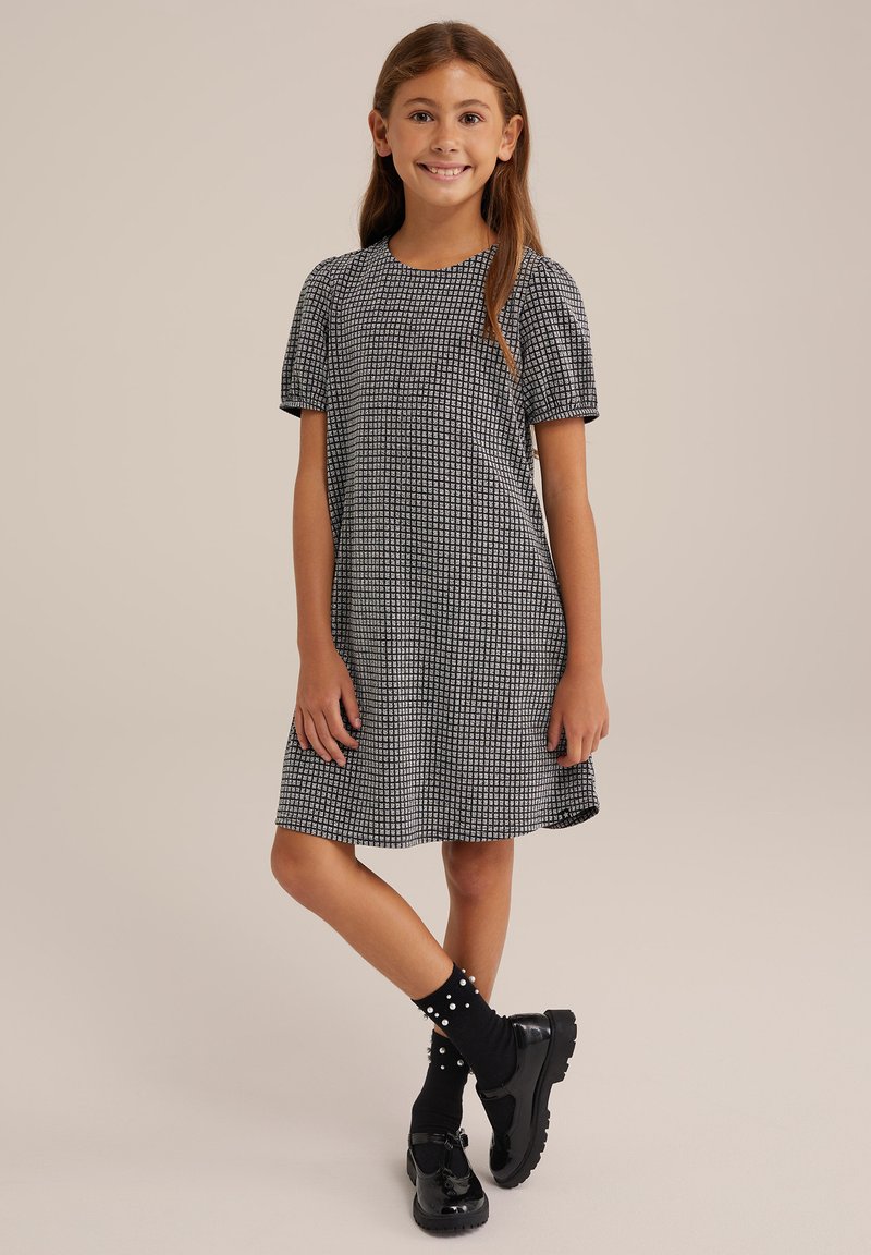 Lyhythihainen mustavalkoinen mekko, jossa on ruudullinen kuvio, pyöreä pääntie ja valuva siluetti, yhdistettynä mustiin kenkiin ja pilkullisiin sukkiin.