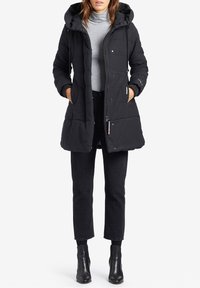 Svart isolerad pufferjacka med huva, med dragkedja framtill, quiltat mönster, sidofickor och ribbade ärmslut. Åtföljs av svarta jeans och stövlar.