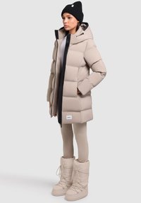 Abrigo acolchado beige con capucha, cremallera negra y parche con logo. Combinado con leggings beige y botas de invierno gruesas de color crema.