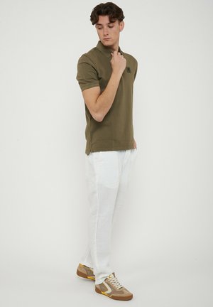 Giovane uomo che indossa una polo verde oliva, pantaloni bianchi e sneakers beige, in piedi e guarda verso il basso con la mano vicino al colletto.