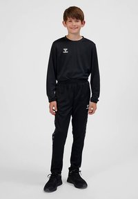 Tuta da atletica nera con maniche lunghe e vita elasticizzata, caratterizzata da un logo sul petto e strisce laterali sui pantaloni, abbinata a scarpe da ginnastica nere.