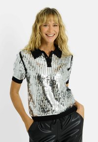 Camisa tipo polo con lentejuelas plateadas, cuello y puños negros, mangas cortas y corte holgado. Textura suave con un acabado brillante.