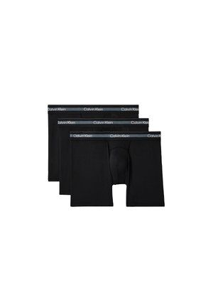 Mężczyzna bez koszulki o muskularnej budowie ciała, ubrany w czarne bokserki Calvin Klein, stojący boso na jednolitym, szarym tle ze skrzyżowanymi rękami.