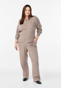 Sieviete stāv, viena roka kabatā, tērpusies bēša krāsas ribota adījuma džemperī ar priekšējām rāvējslēdzēju un atbilstošās platām bikšu biksēm, melnās apavos ar naglām.