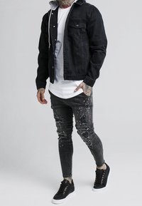 Schwarze Jeansjacke über einem weißen T-Shirt, kombiniert mit verwaschenen schwarzen Jeans mit Farbspritzern und schwarzen Sneakers mit weißen Sohlen.